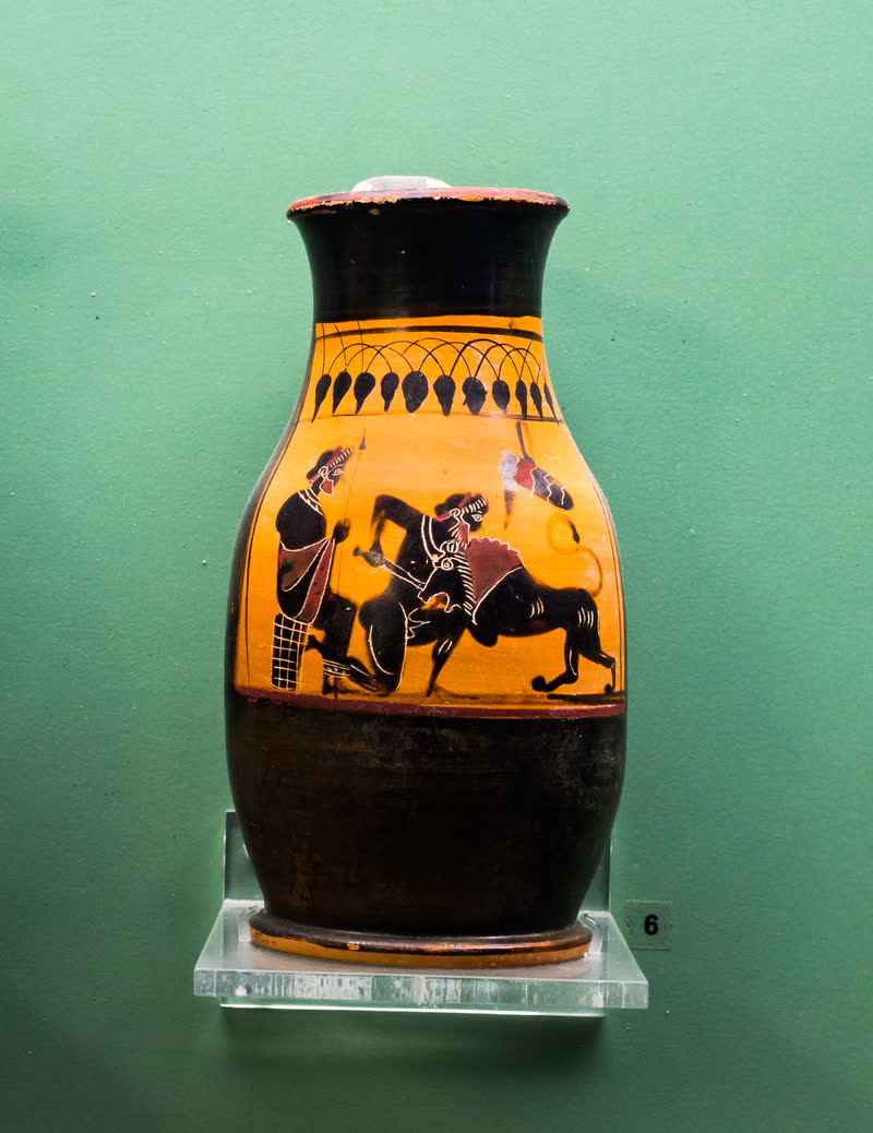 Héracles crava a espada no Leão de Nemeia, que se ergue contra ele; à esquerda, Iolau sentado num bloco, segurando a lança, e a aljava pendurada ao alto. Olpe ática de figuras negras, ca. 530–520 a.C., da Classe do Louvre F 158; achada na tumba 19 de Marmaro, em Ialisos. Museu Arqueológico de Rodes (15443).